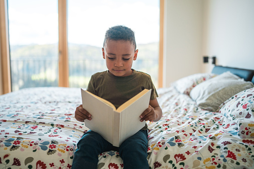 Adorable Mixed race preschooler boy sitting on a bed in a domestic bed room reading a book.
