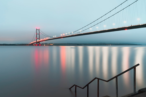 The Humber bridge lit up by bright colorful lights reflecting on the estuary just dawn breaks in Hessle, Yorkshire, UK.