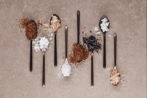 Different kinds of salts in a spoonfuls, top view. Composition with different salt  on a beige background. Sea salt, cooking salt, Himalayan and black salt .