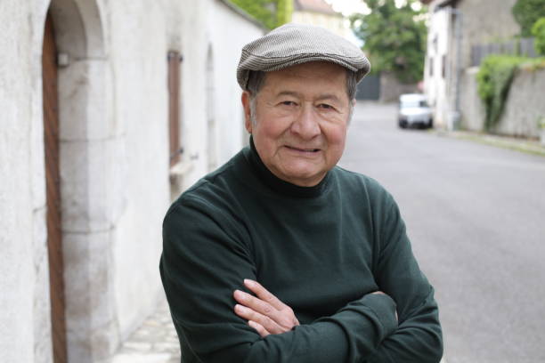 A senior ethnic businessman in his seventies is posing with his arms crossed. He wears an elegant green turtle neck sweater and a hat.