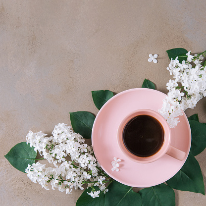 High angle view of black coffee in pastel pink cup surrounded with green leaves and white lilac flowers on rustic brown stone background with copy space.