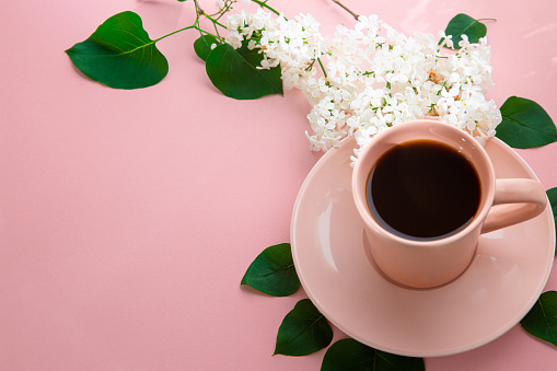 Black coffee in pastel pink cup surrounded with green leaves and white lilac flowers over pink background with copy space.