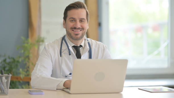 istock Male Doctor Smiling at Camera while Working on Laptop 1493543874
