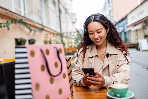 Smiling young woman looking at smart phone in cafe shop after shopping