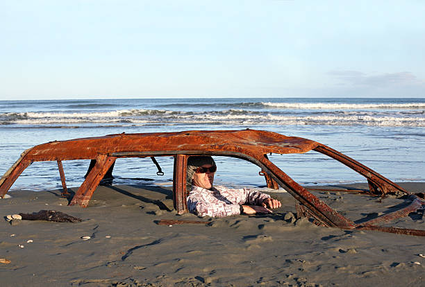 Man sitting in rusty car buried on a beach.