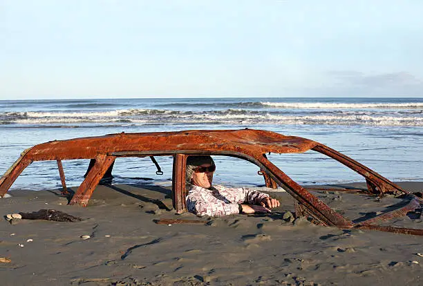 Photo of Man sitting in rusty car buried on a beach.