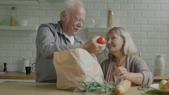 Senior man bringing bag with groceries to wife and unpacking food products in kitchen at home