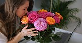 Young beautiful lady enjoys aroma of colorful bouquet of rose flowers. Romantic scents and atmosphere at home, vase stands on desk with laptop