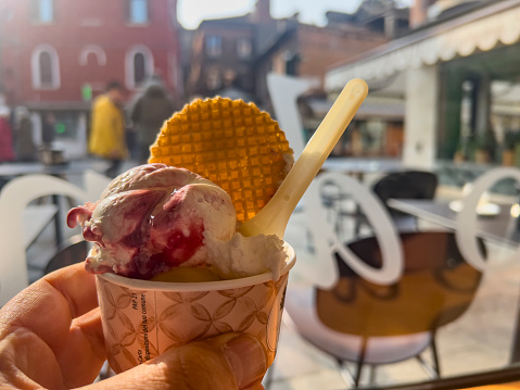 Italian food background: Hand holding beautiful arranged traditional ice cream with cherry souse, crispy waffle and a spoon under sunlight inside a restaurant with window view from an Gelateria in Venice.