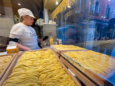 Venice - Italy, 31st January 2023: Italian culture and food background, editorial with copy space. Female person with white head and apron preparing pasta from  fresh dough in front of a restaurant window.