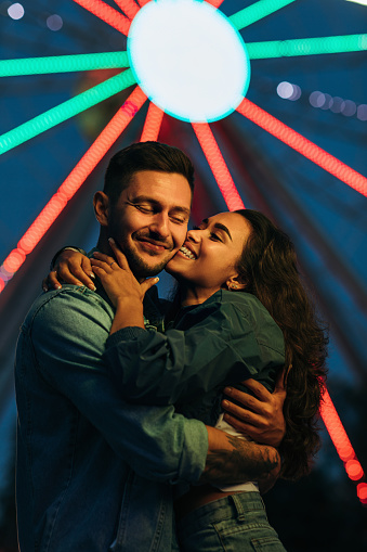 Young happy couple against Ferris wheel during the festival. Woman kissing her smiling boyfriend at night.