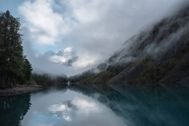 istock Tranquil scenery with snow castle in clouds. Mountain creek flows from forest hills into glacial lake. Snowy mountains in fog clearance, small river and coniferous trees reflected in calm alpine lake. 1493623972