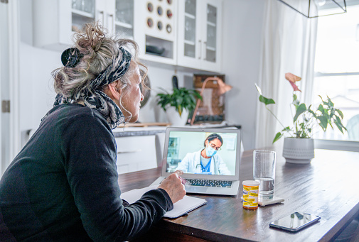 A senior woman sits at the kitchen table in the comfort of her own home, with a laptop open in front of her.  She is dressed casually and smiling as she talks virtually with her doctor about her medication.