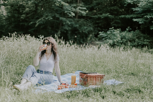 A beautiful young caucasian brunette girl in jeans, a t-shirt and sunglasses sits on a bedspread with a wicker basket, food and drinks orange juice from a glass in a public park, close-up side view. Outdoor picnic concept.