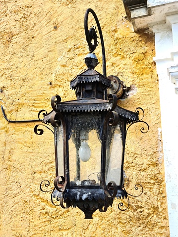 Very old outdoor lamp on a yellow wall during daylight hours.