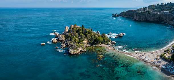 Isola Bella Islet in the bay of famous Taormina on Sicily Island. Tourists enjoying their beach vacation on the small white sandbar and coastal beach from Taormina towards Isola Bella. Stitched Drone Point of View Panorama. Isola Bella, Taormina, Messina, Sicily, Italy, Southern Europe, Europe.