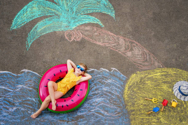 glückliches kleines vorschulmädchen im badeanzug auf aufblasbarem ring mit meer, sand, palme mit bunten kreiden auf asphalt gemalt. süßes kind mit spaß mit kreidebild. sommer, urlaub, sommer - romrodinka stock-fotos und bilder