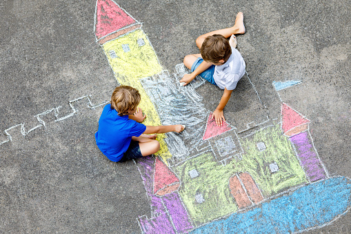Two little kids boys drawing knight castle with colorful chalks on asphalt. Happy siblings and friends having fun with creating chalk picture and painting. Creative leisure for children in summer