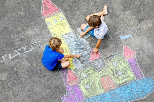 Dois meninos dos miúdos pequenos que desenham o castelo do cavaleiro com giz coloridos no asfalto. Irmãos felizes e amigos que têm o divertimento com criação do retrato e da pintura do giz. Lazer criativo para crianças no verão. - foto de acervo