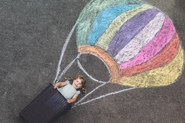 glückliches kleines kleinkind mädchen fliegen in heißluftballon mit bunten kreiden in regenbogenfarben auf dem boden oder asphalt im sommer gemalt. nettes kind mit spaß. kreative freizeit für kinder. - romrodinka stock-fotos und bilder