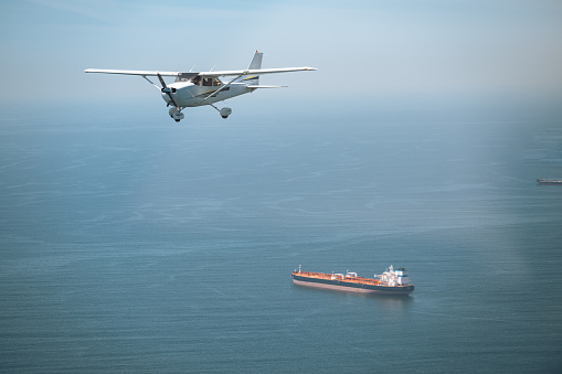 A light aircraft soars over the expanse of the ocean, with cargo ships visible below, creating a captivating juxtaposition of air and sea.