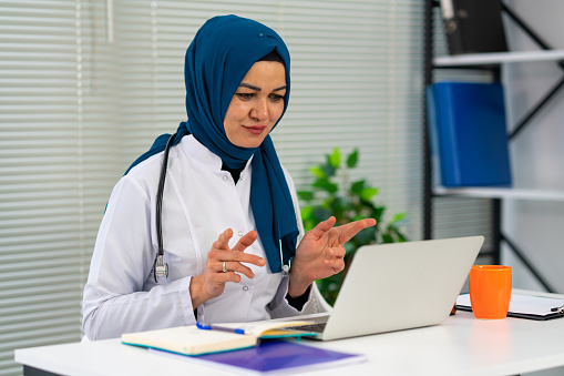 Doctor online consultation with patient, muslim woman in hijab working remotely with laptop for video call, consulting patients sitting inside modern clinic office