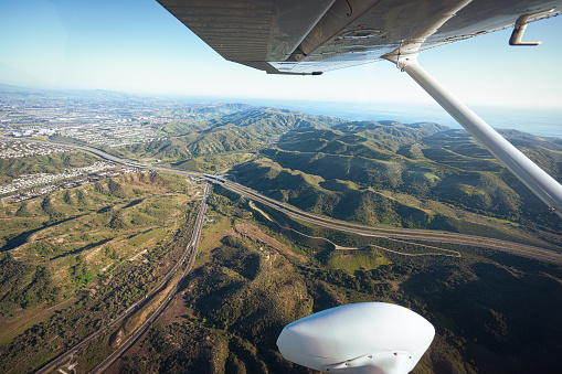 Through the airplane window, as the camera lens captures the scene below, the sprawling landscape of California comes into view, with its diverse terrain, vibrant cities, and scenic horizon stretching as far as the eye can see.