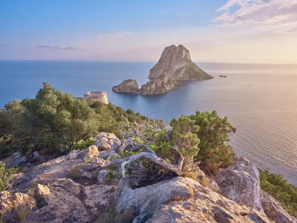 Wide-angle view of the rocky isles of Es Vedrà and Es Vedranell, off the south-western coast of Ibiza, and the 18th-century Torre des Savinar (also known as Torre del Pirata). Picturesque clouds, calm waters reflecting the oblique, golden light of a summer sunset, steep cliffs covered with lush Mediterranean scrub. High level of detail, natural rendition, realistic feel. Developed from RAW.