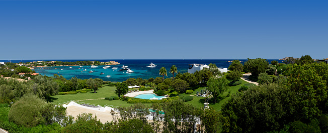 Escape to Paradise: Behold the captivating panorama of Porto Cervo village nestled in the picturesque Sardinia, Sardinia, Italy. This enchanting view showcases a magnificent villa surrounded by lush gardens and a dazzling swimming pool. Beyond, the sparkling bay glimmers with luxurious yachts, while the turquoise and deep blue sea invites you to dive into its crystal-clear depths. As the sun illuminates the landscape, the clear sky completes this postcard-perfect scene. Immerse yourself in the beauty of Porto Cervo, where opulence meets natural splendor.