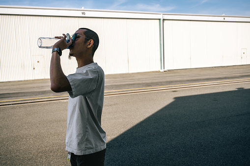 A young man stands in front of the hangar, taking a refreshing sip of water, rejuvenating himself amidst the aviation setting.