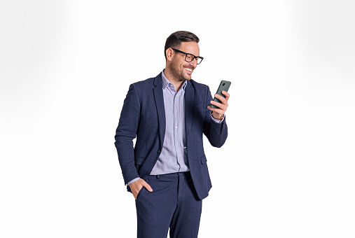 Handsome male professional manager with hand in pocket messaging online on mobile phone. Elegant businessman reading e-mails on cellphone while standing isolated over white background