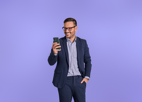 Smiling male professional chief with hand in pocket checking messages over smart phone. Young businessman dressed in formals using social media apps on cellphone isolated against blue background