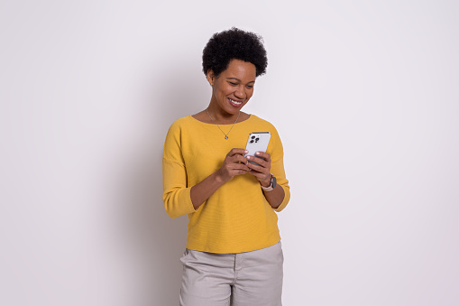 Smiling young woman with short afro hair text messaging on smart phone. Beautiful female dressing in casuals using social media over cellphone while standing isolated on white background