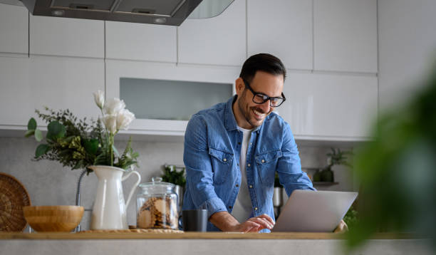 istock Cheerful young businessman dressed in denim shirt sending reports over laptop while standing at kitchen island. Happy male entrepreneur surfing net over computer while working from home office 1493705679