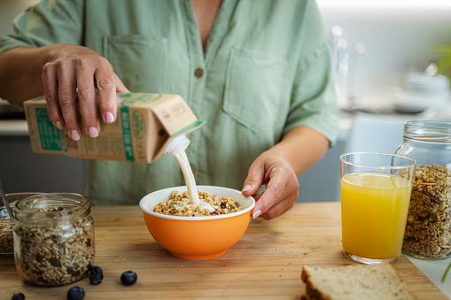 Cropped shot of woman's hands pouring milk from a carton package into a bowl full of healthy breakfast cereal on kitchen counter