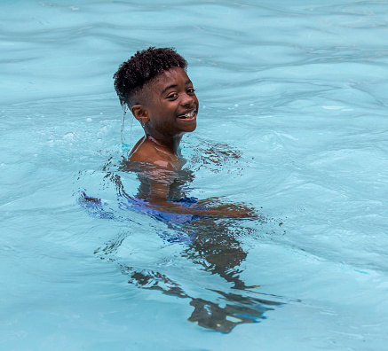 African-American boy, having a blast smiling, and laughing ￼ as he plays in the swimming pool, dipping under and coming out as a water rows ￼off of him in different shapes and forms. ￼