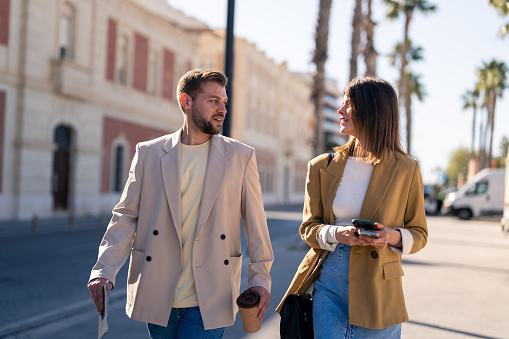 Young beautiful stylish woman and handsome man in beige blazer meeting and walking down the city street on a sunny day.