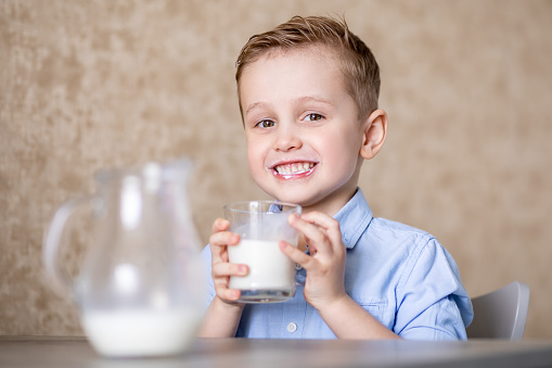A little boy in a blue shirt is drinking milk from a glass mug with pleasure. The baby is smiling. In the foreground is a blurred carafe background. Emotional reaction.