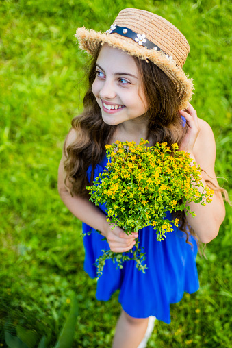 Cute smiling girl wearing straw hat collecting yellow flowers in field.