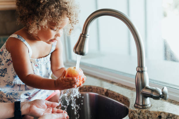 Cute toddler with mom together at home washing apples in the sink, domestic life stock photo