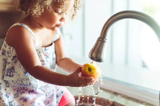 Photo of Cute toddler at home washing apples in the sink, domestic life