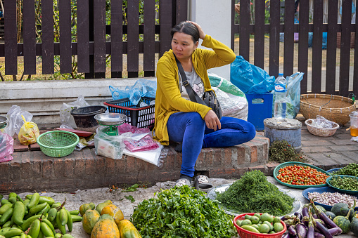 Morning market, Luang Prabang, Laos - March 15th 2023: Female greengrocer waiting for customers in her shop on the ground at the famous morning food market in Luang Prabang which used to be the capital on Laos