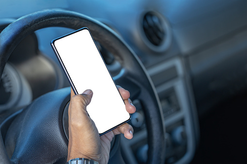 Detail of the hand of a driver holding a mobile phone with a blank screen on the steering wheel of the car with copy space.