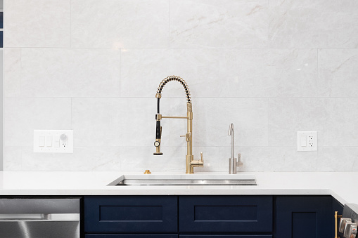 A kitchen sink detail shot in a white and blue kitchen with a gold faucet, marble countertop, and marble tile backsplash.