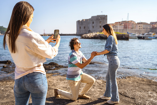Man proposing to his girlfriend by the sea edge on a beautiful sunny day while their friend is taking pictures