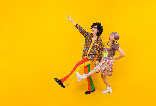 Pareja hippie asiática se viste de moda vintage de los 80 con colorida ropa retro mientras bailan juntos aislados sobre fondo amarillo para una fiesta de atuendo elegante y un concepto de cultura pop photo