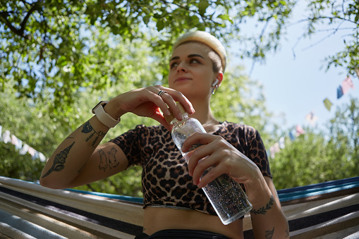 Beautiful diverse female person with short hair drinking water from a glass bottle in a sunny summer park