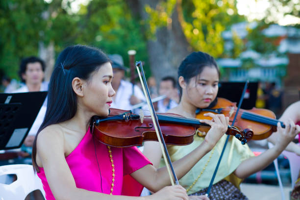 Two thai women are playing violin. They are members of small orchestra exercising for evening event in public. Scene is at History week in Phitsanulok with traditional food, clothings and performances about history and battles with Myanmar. Performance takes place at historical grounds of Wat Wihan Thong Historical Site near Nan river and is local way of keeping history alive