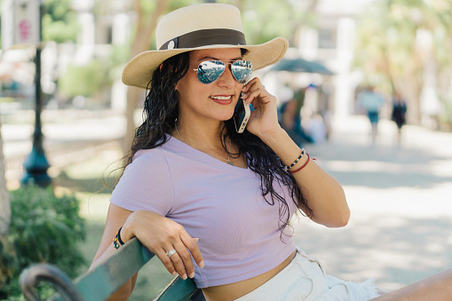 Latin woman sitting on a park bench talking on the phone, she wears a hat and sunglasses and is smiling.