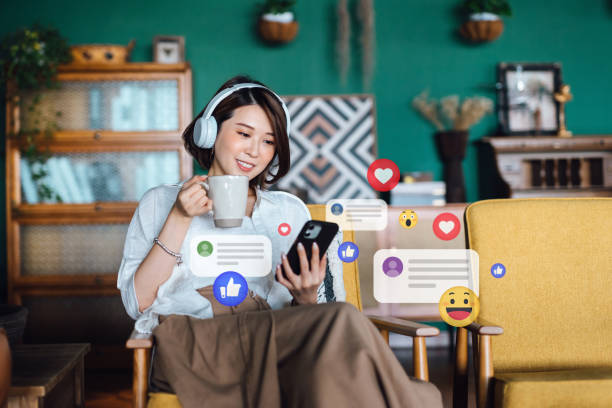 belle jeune femme asiatique avec des écouteurs se relaxant à la maison et utilisant un smartphone, vérifiant les médias sociaux sur le téléphone portable, reçoit des notifications, des likes, des vues et des commentaires. mode de vie des jeunes, mé - audio branding photos et images de collection